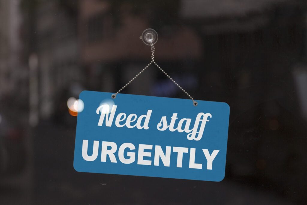 Close-up on a red and white sign in a window with written the message - Need staff urgently